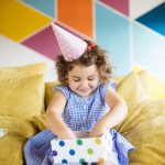 Cute little girl with dark curly hair in birthday cap happily op