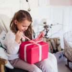 Little girl holding a Christmas gift box. Cute kid sitting on the chair and unpacks xmas presents