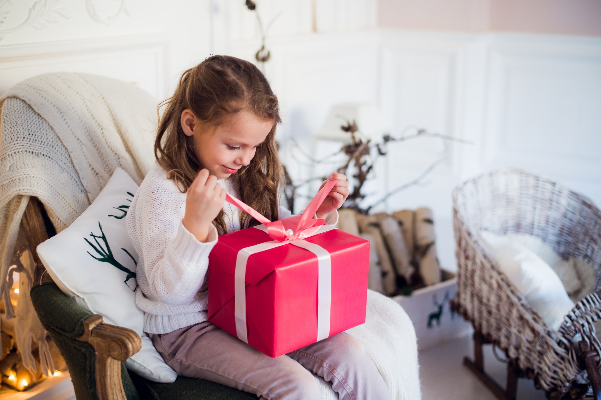 Little girl holding a Christmas gift box. Cute kid sitting on the chair and unpacks xmas presents