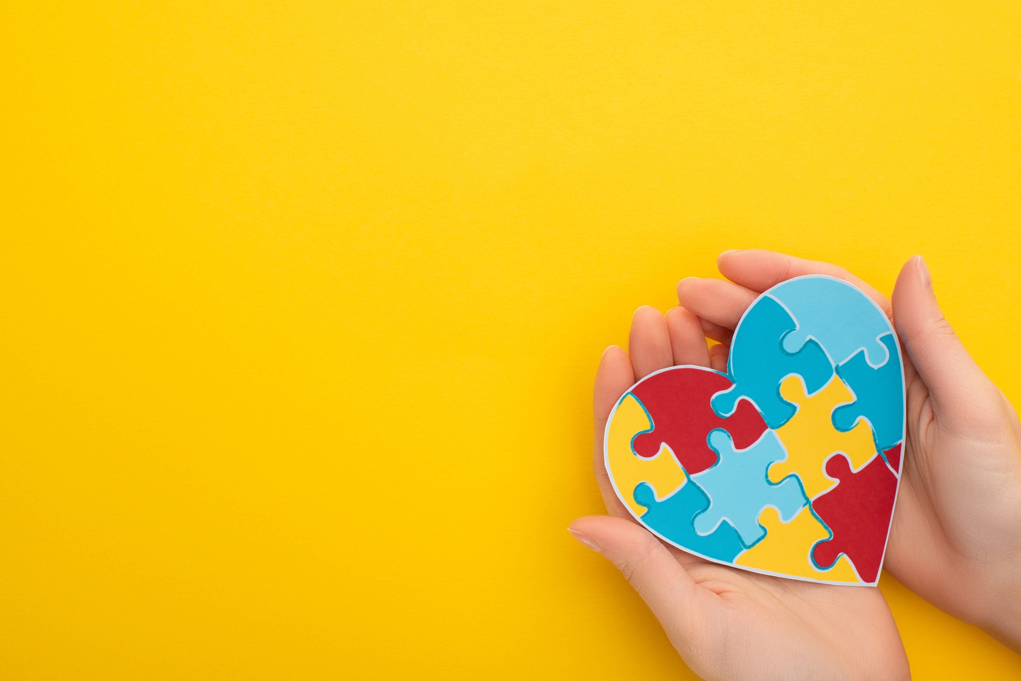 partial view of woman holding colorful puzzle heart on yellow for World Autism Awareness Day
