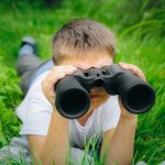 Boy looking through binoculars in green grass plants birdwatching nature exploration kid adventure