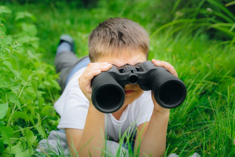 Boy looking through binoculars in green grass plants birdwatching nature exploration kid adventure