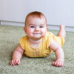 Portrait of smiling little Caucasian baby girl in yellow dress. Child trying to crawl on a floor.