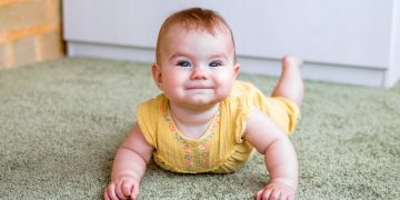 Portrait of smiling little Caucasian baby girl in yellow dress. Child trying to crawl on a floor.