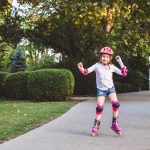 Little girl rides on rollers in summer park. Child wearing protection pads and safety helmet